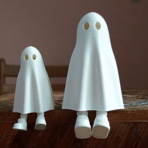 May include: Two white ghost figurines with black eyes, sitting on a wooden surface. The ghost on the left is smaller than the ghost on the right.