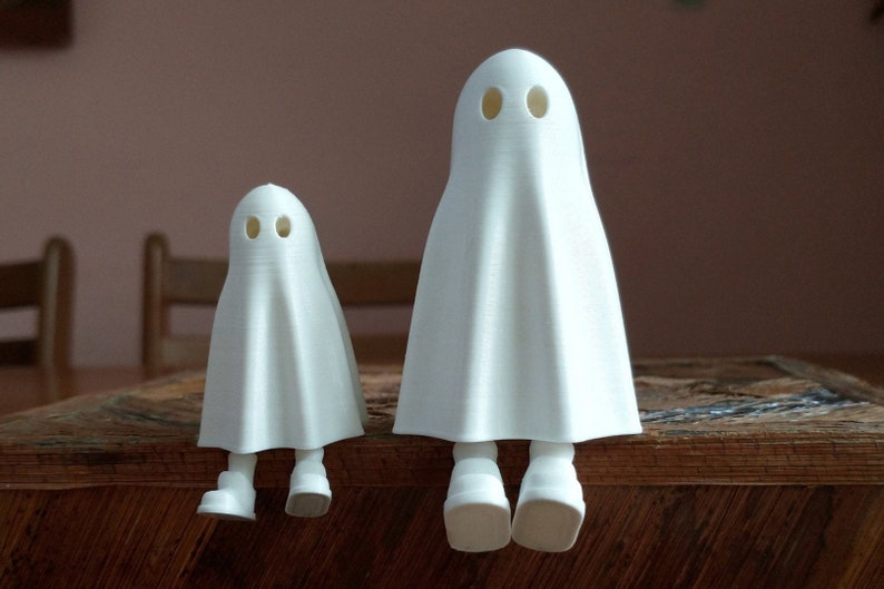 May include: Two white ghost figurines with black eyes, sitting on a wooden surface. The ghost on the left is smaller than the ghost on the right.