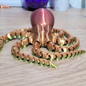 May include: A 3D printed octopus figurine with a copper-coloured body and green tentacles. The octopus is sitting on a wooden surface.