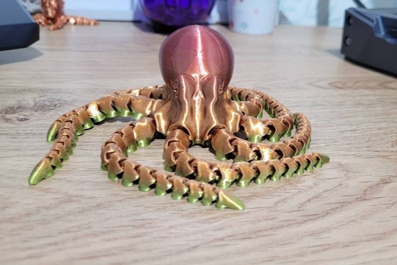 May include: A 3D printed octopus figurine with a copper-coloured body and green tentacles. The octopus is sitting on a wooden surface.