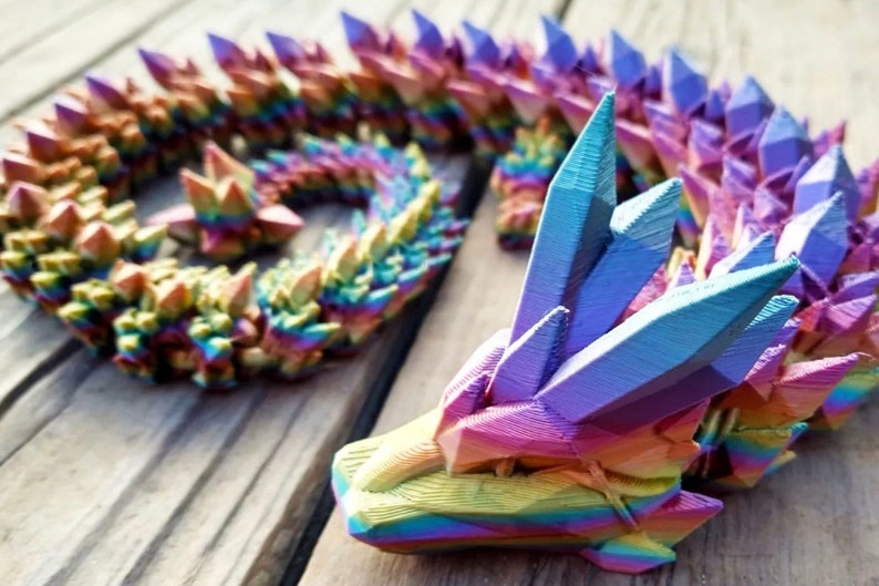 May include: A rainbow-colored, 3D printed dragon sculpture with a crystal-like texture. The dragon is curled up in a coil shape, with its head raised and its eyes closed.