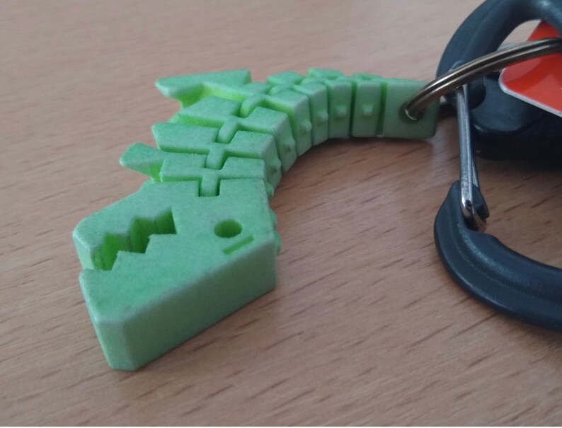 Puede incluir: Un llavero de pl&aacute;stico verde en forma de esqueleto de dinosaurio. El dinosaurio est&aacute; en posici&oacute;n enroscada con la cabeza mirando hacia el lado izquierdo de la imagen. El llavero est&aacute; unido a un mosquet&oacute;n negro.