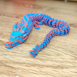 Puede incluir: Una figurita de drag&oacute;n impresa en 3D con rayas azules y rojas. El drag&oacute;n est&aacute; en posici&oacute;n enroscada con la cabeza levantada.