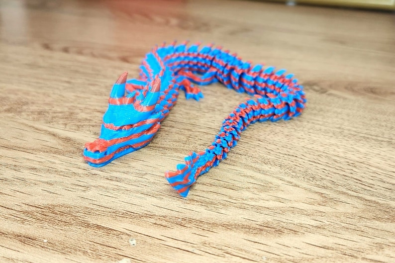 Puede incluir: Una figurita de drag&oacute;n impresa en 3D con rayas azules y rojas. El drag&oacute;n est&aacute; en posici&oacute;n enroscada con la cabeza levantada.