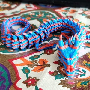 Puede incluir: Una figurita de drag&oacute;n impresa en 3D azul y roja con una espalda espinosa y una cara detallada.