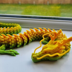 May include: A 3D printed dragon figurine with a green and yellow gradient color scheme. The dragon is curled up in a resting position with its head resting on its body. The dragon has a detailed, scaly texture and a long, flowing tail.