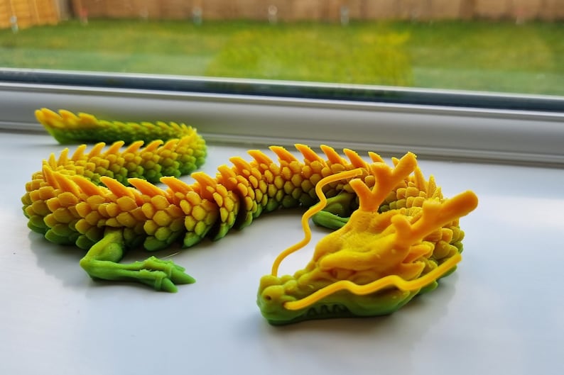 May include: A 3D printed dragon figurine with a green and yellow gradient color scheme. The dragon is curled up in a resting position with its head resting on its body. The dragon has a detailed, scaly texture and a long, flowing tail.