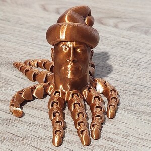 Puede incluir: Una figurita impresa en 3D de una cabeza humana con un gorro de Pap&aacute; Noel y un cuerpo de pulpo. La figurita est&aacute; hecha de un material de color cobre.