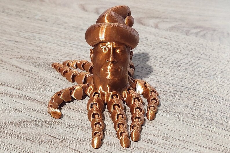 Puede incluir: Una figurita impresa en 3D de una cabeza humana con un gorro de Pap&aacute; Noel y un cuerpo de pulpo. La figurita est&aacute; hecha de un material de color cobre.