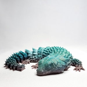May include: A 3D printed model of a blue and green lizard with a spiky back. The lizard is curled up in a defensive position with its head tucked in.