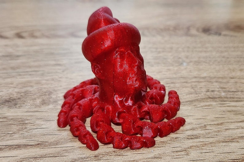 Puede incluir: Una figura de pulpo roja impresa en 3D con una superficie texturizada. El pulpo tiene una cabeza redonda y ocho tent&aacute;culos.