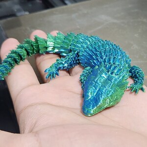 May include: A 3D printed model of a lizard with a blue and green iridescent finish. The lizard is posed in a curled position with its tail wrapped around its body.