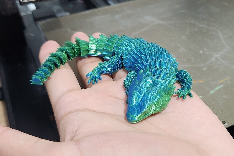 May include: A 3D printed model of a lizard with a blue and green iridescent finish. The lizard is posed in a curled position with its tail wrapped around its body.