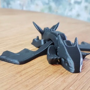 May include: A black 3D printed dragon figurine with wings outstretched. The dragon has a detailed head with sharp teeth and horns.