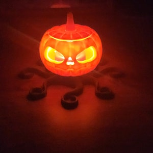 May include: A glowing orange jack-o'-lantern with a scary face sits on a black, octopus-shaped stand.