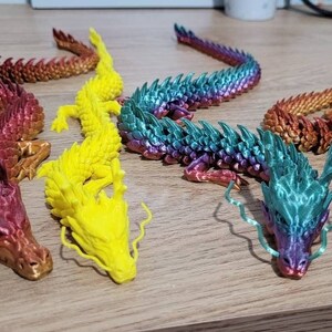May include: Five colorful 3D printed dragon figurines. The dragons are in various poses and have detailed scales. The dragons are red, yellow, green, blue, and purple.