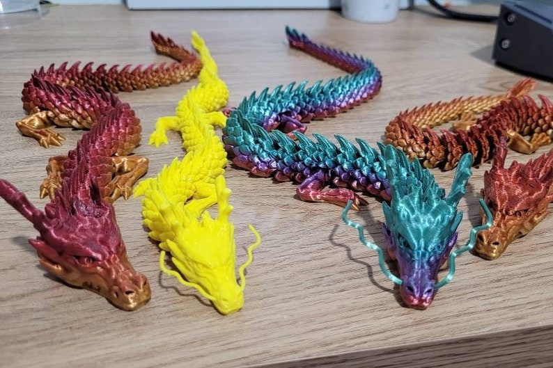 May include: Five colorful 3D printed dragon figurines. The dragons are in various poses and have detailed scales. The dragons are red, yellow, green, blue, and purple.