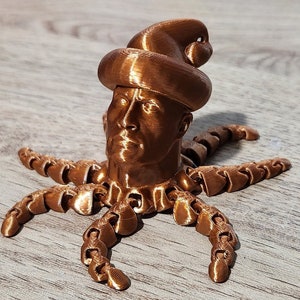Puede incluir: Una escultura impresa en 3D de una cabeza humana con un sombrero, con un acabado de cobre. La cabeza est&aacute; unida a un cuerpo de pulpo de m&uacute;ltiples patas.