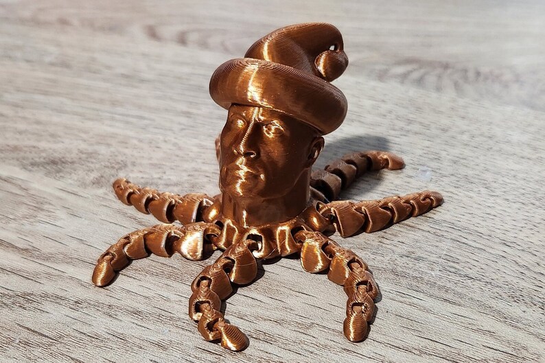 Puede incluir: Una escultura impresa en 3D de una cabeza humana con un sombrero, con un acabado de cobre. La cabeza est&aacute; unida a un cuerpo de pulpo de m&uacute;ltiples patas.