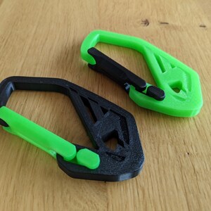 May include: Two 3D-printed carabiner clips. One is black with a green clasp, and the other is green with a black clasp. The clips have a geometric design and are lying on a wooden surface.
