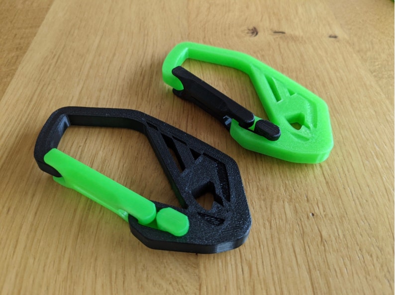 May include: Two 3D-printed carabiner clips. One is black with a green clasp, and the other is green with a black clasp. The clips have a geometric design and are lying on a wooden surface.