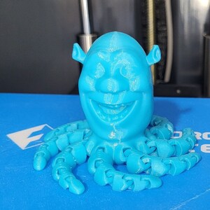 Puede incluir: Una figura de Shrek de color azul impresa en 3D con un cuerpo de pulpo. La figura tiene una gran sonrisa y est&aacute; impresa en azul claro.
