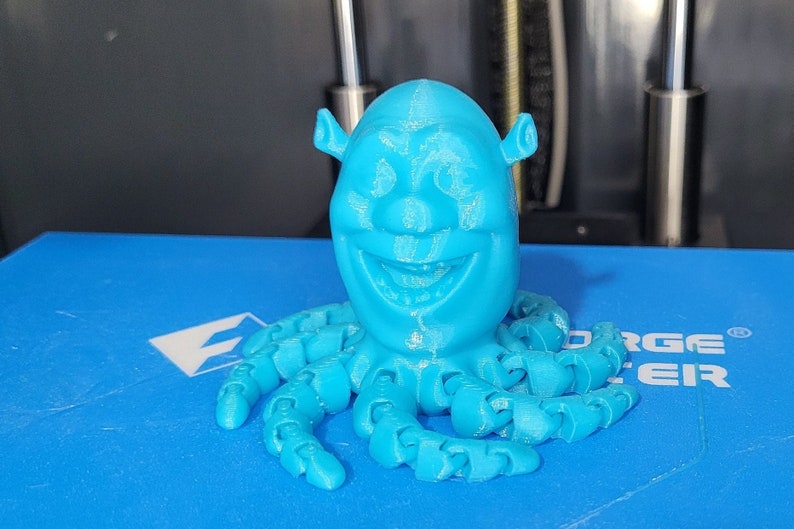 Puede incluir: Una figura de Shrek de color azul impresa en 3D con un cuerpo de pulpo. La figura tiene una gran sonrisa y est&aacute; impresa en azul claro.