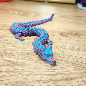 Puede incluir: Una figura de drag&oacute;n impresa en 3D de color azul y rojo con una espalda y cola espinosa. El drag&oacute;n est&aacute; en posici&oacute;n con la cabeza levantada y la boca abierta.