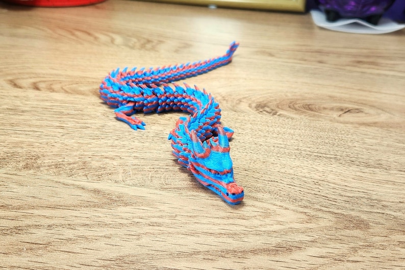 Puede incluir: Una figura de drag&oacute;n impresa en 3D de color azul y rojo con una espalda y cola espinosa. El drag&oacute;n est&aacute; en posici&oacute;n con la cabeza levantada y la boca abierta.