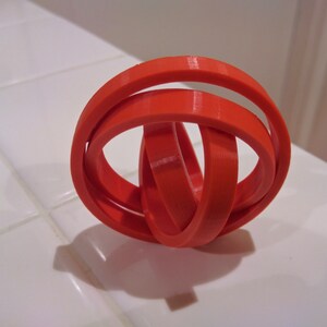 Puede incluir: Una escultura de anillos entrelazados de color naranja brillante, impresa en 3D. Los anillos est&aacute;n entrelazados y crean un patr&oacute;n geom&eacute;trico complejo.