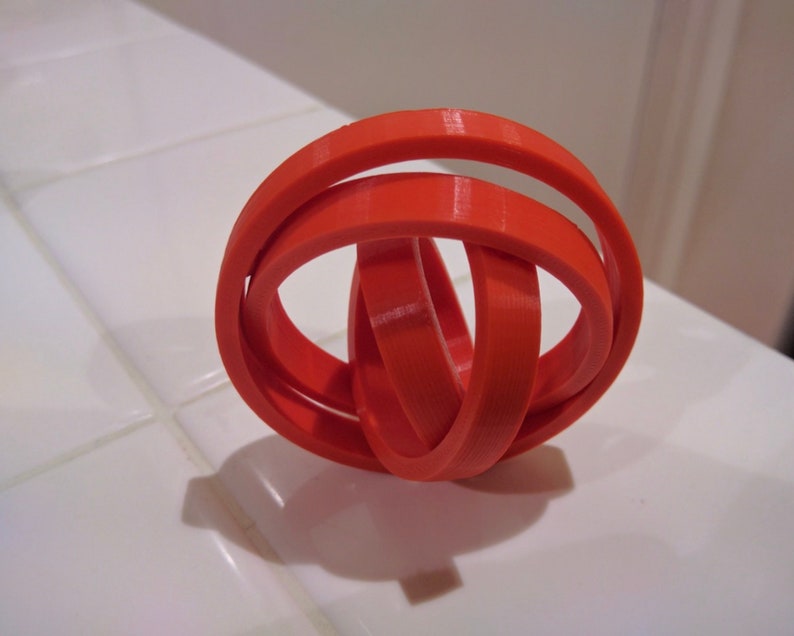 Puede incluir: Una escultura de anillos entrelazados de color naranja brillante, impresa en 3D. Los anillos est&aacute;n entrelazados y crean un patr&oacute;n geom&eacute;trico complejo.