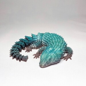 May include: A 3D printed model of a blue and green lizard with a segmented body. The lizard has a textured, scaly appearance and is posed with its head down.