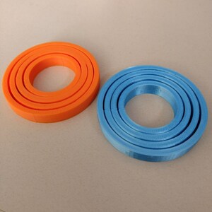 Puede incluir: Dos juegos de anillos conc&eacute;ntricos, uno naranja y otro azul. Los anillos est&aacute;n hechos de un material liso, similar al pl&aacute;stico, y est&aacute;n apilados uno encima del otro.