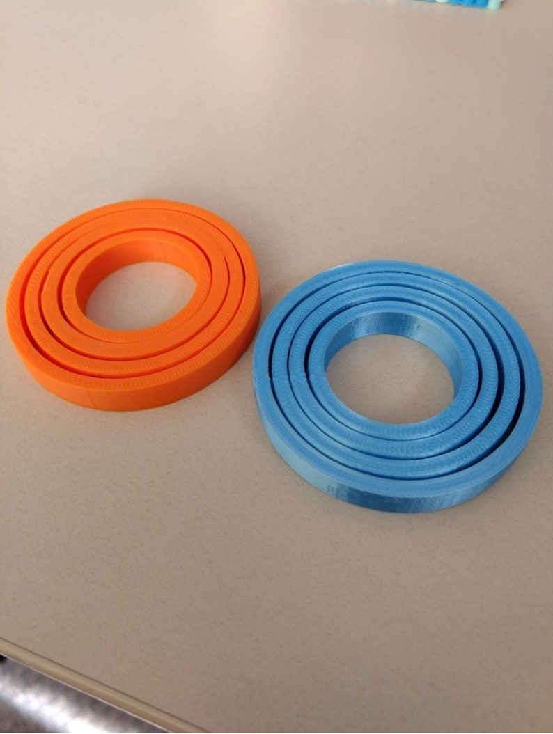 Puede incluir: Dos juegos de anillos conc&eacute;ntricos, uno naranja y otro azul. Los anillos est&aacute;n hechos de un material liso, similar al pl&aacute;stico, y est&aacute;n apilados uno encima del otro.