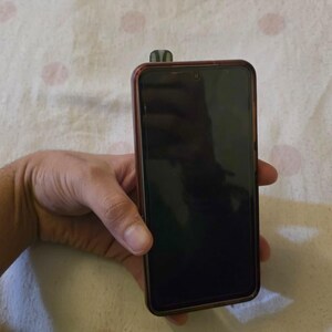 K&ouml;nnte beinhalten: Ein schwarzes Smartphone mit dunklem Bildschirm und braunem Rahmen wird in einer Hand gehalten. Ein kleines, dunkel gef&auml;rbtes Objekt ist oben am Telefon befestigt. Der Hintergrund ist ein wei&szlig;er Stoff mit rosa Punkten.