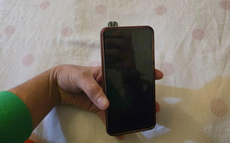 K&ouml;nnte beinhalten: Ein schwarzes Smartphone mit dunklem Bildschirm und braunem Rahmen wird in einer Hand gehalten. Ein kleines, dunkel gef&auml;rbtes Objekt ist oben am Telefon befestigt. Der Hintergrund ist ein wei&szlig;er Stoff mit rosa Punkten.
