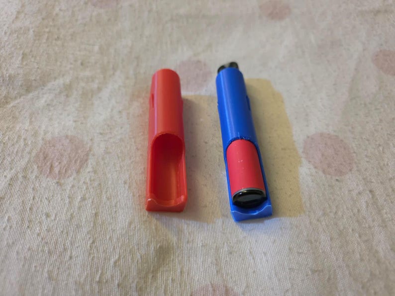 K&ouml;nnte beinhalten: Ein rotes und blaues Feuerzeuggeh&auml;use mit einem Feuerzeug darin. Das Geh&auml;use besteht aus Kunststoff und hat eine rechteckige Form. Das Feuerzeug ist rot und schwarz. Das Geh&auml;use ist offen und das Feuerzeug ist sichtbar.
