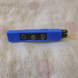 K&ouml;nnte beinhalten: Ein blaues elektronisches Ger&auml;t mit einem schwarzen rechteckigen Abschnitt mit drei silbernen Kreisen. Das Ger&auml;t hat eine rechteckige Form mit einem abgerundeten Ende und einer dunkelgr&uuml;nen Spitze. Der Hintergrund ist ein wei&szlig;er Stoff mit rosa Flecken.