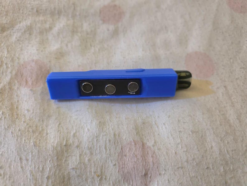 K&ouml;nnte beinhalten: Ein blaues elektronisches Ger&auml;t mit einem schwarzen rechteckigen Abschnitt mit drei silbernen Kreisen. Das Ger&auml;t hat eine rechteckige Form mit einem abgerundeten Ende und einer dunkelgr&uuml;nen Spitze. Der Hintergrund ist ein wei&szlig;er Stoff mit rosa Flecken.