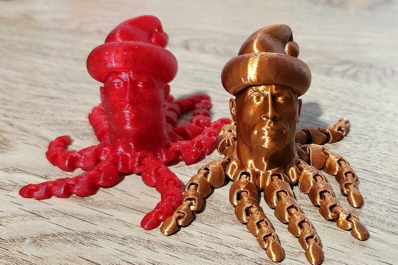 Puede incluir: Dos figuritas de pulpo impresas en 3D con cabezas humanas que llevan gorros de Pap&aacute; Noel. Un pulpo es rojo y el otro es cobre.