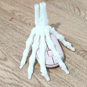 May include: A white plastic skeleton hand resting on top of a white computer mouse. The mouse is on a light brown wooden surface.
