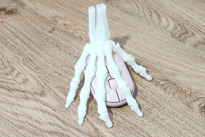 May include: A white plastic skeleton hand resting on top of a white computer mouse. The mouse is on a light brown wooden surface.