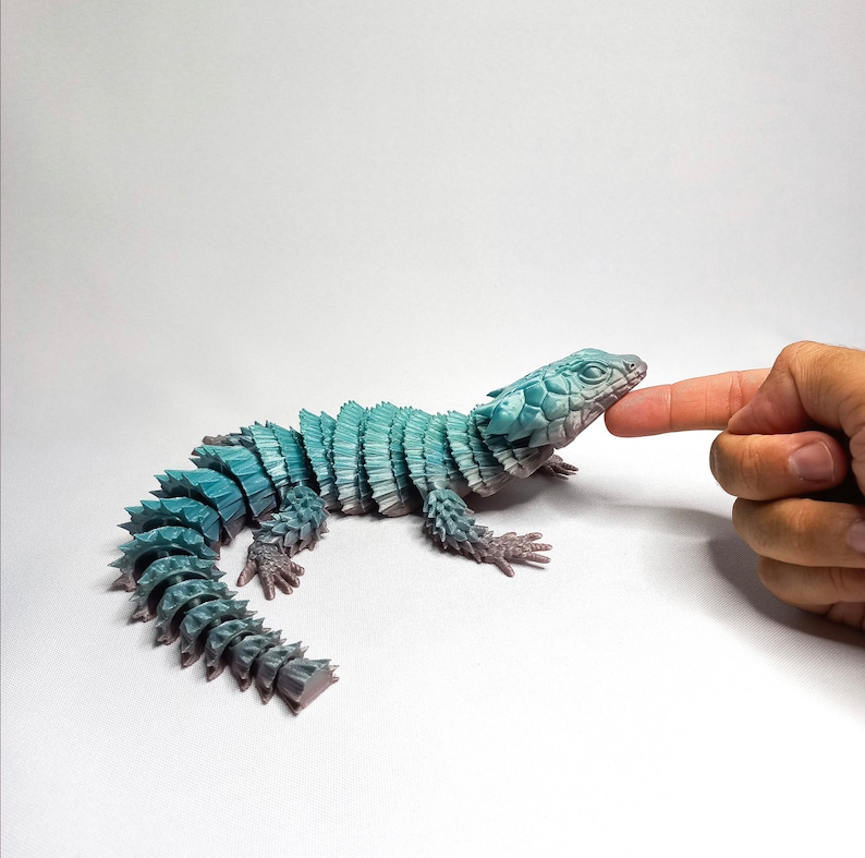 May include: A blue and gray 3D printed model of a lizard with a spiky tail and a textured body. The lizard is posed as if it is walking on a white surface. A hand is reaching out to touch the lizard's head.