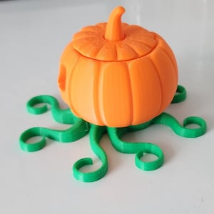 May include: A 3D printed orange pumpkin with a lid, sitting on a green, 3D printed base that resembles a vine.