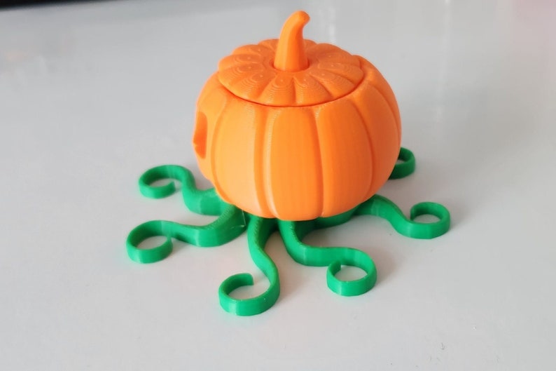 May include: A 3D printed orange pumpkin with a lid, sitting on a green, 3D printed base that resembles a vine.