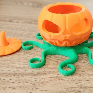 May include: Orange pumpkin with a carved face and a green octopus base. The pumpkin has a removable lid.