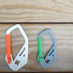 May include: Two 3D-printed carabiners, one white with an orange gate and the other gray with a green gate, are displayed against a wood grain background. The text "ddf3d.com" and "selflex" are visible.