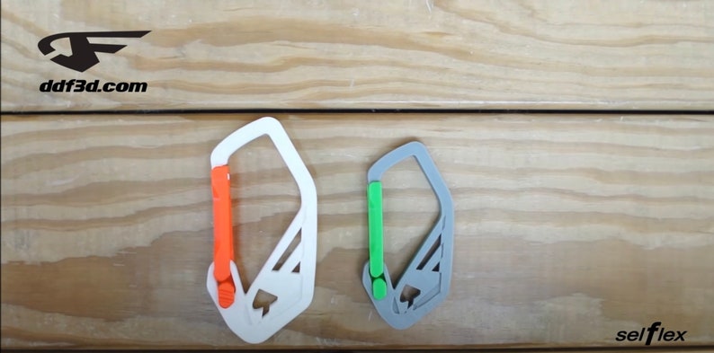 May include: Two 3D-printed carabiners, one white with an orange gate and the other gray with a green gate, are displayed against a wood grain background. The text "ddf3d.com" and "selflex" are visible.