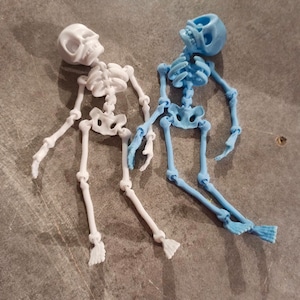 May include: Two articulated skeleton figurines, one white and one light blue, lying on a gray surface. The skeletons are posed in a relaxed, almost playful manner.