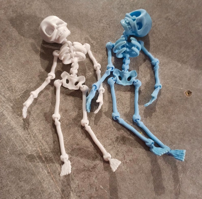 May include: Two articulated skeleton figurines, one white and one light blue, lying on a gray surface. The skeletons are posed in a relaxed, almost playful manner.